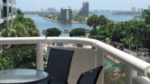Ein Balkon mit Glastisch und Stühlen, der auf ein Gewässer und eine Stadtansicht blickt.