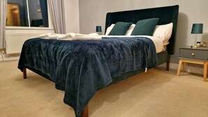 A neatly made bed with a dark blue blanket, a white duvet, and green pillows. There is a small bedside table with a lamp and a nightstand with a lamp. The room has beige carpet and a window with curtains. - Stunning Luxury 2 Bed City Centre Apartment (Derby)