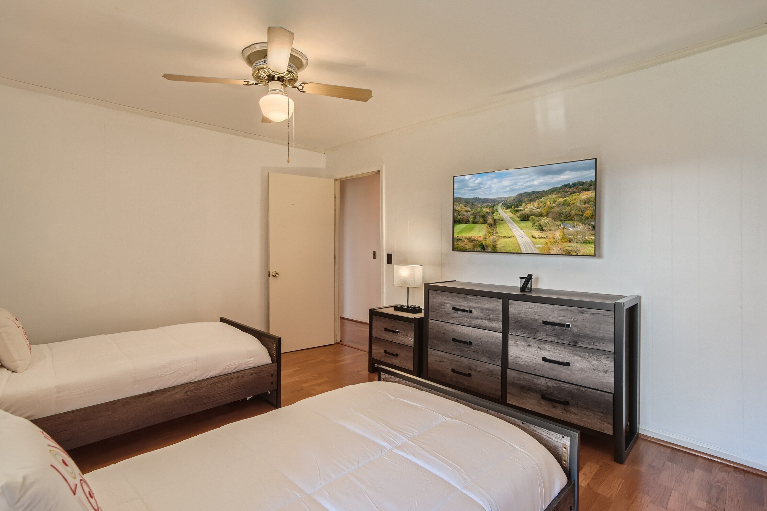 Un dormitorio con dos camas individuales, un aparador de madera, un ventilador de techo y un cuadro con un paisaje en la pared.