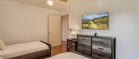 Un dormitorio con dos camas individuales, un aparador de madera, un ventilador de techo y un cuadro con un paisaje en la pared.