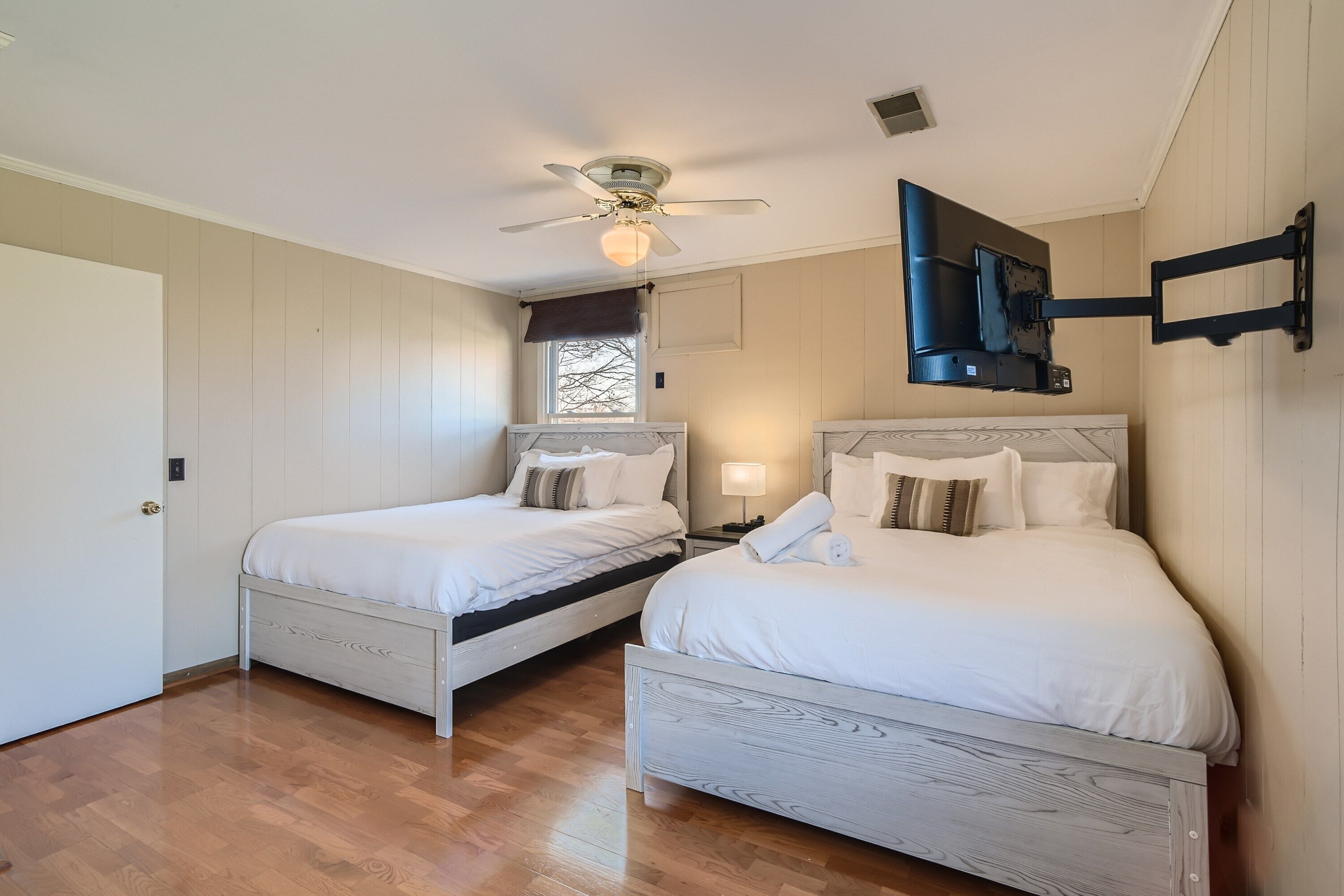 A bedroom with two beds, a ceiling fan, a mounted TV, and a window with a view.