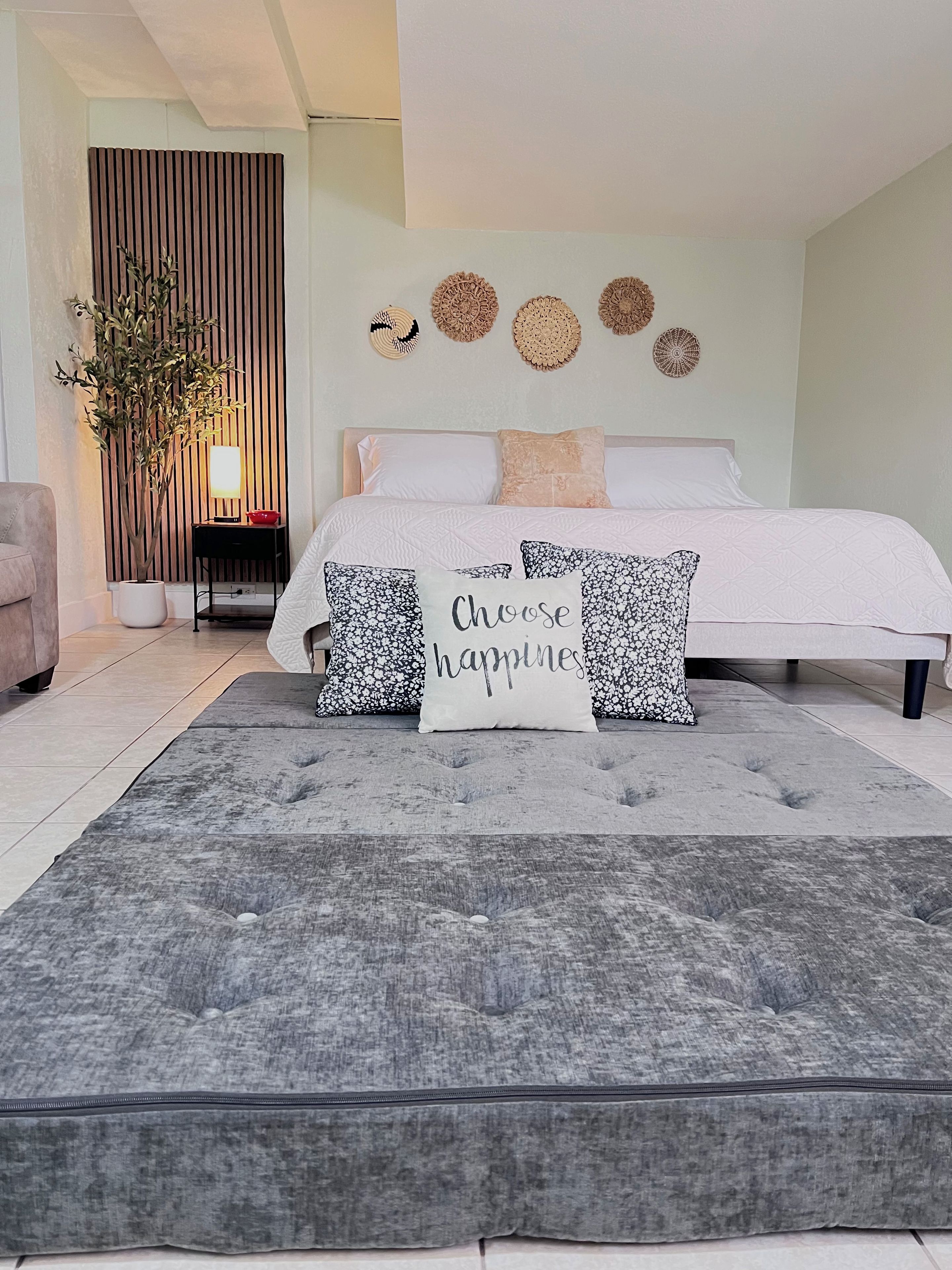 A bedroom with a bed, pillows, a bedside table, a lamp, a potted plant, and decorative wall art.