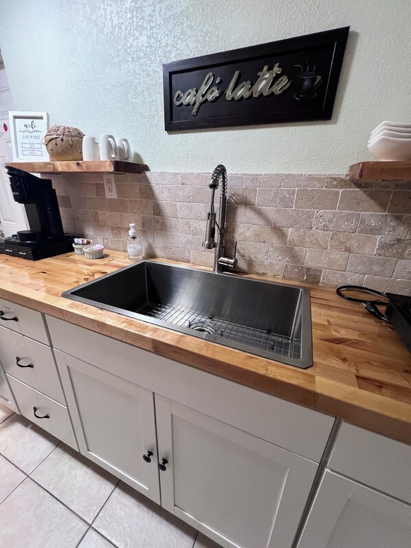 Una cocina con un lavamanos de acero inoxidable, encimera de madera, armarios blancos, una cafetera y un letrero en la pared que dice 'café latte'.