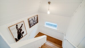 A staircase with white walls and wooden steps leading to a landing. There are two framed pictures on the wall, one of a donkey and another of a horse. A small window with a view of the outside is visible on the landing.