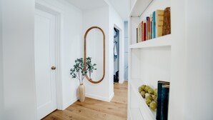 A hallway with white walls, a wooden floor, a door, a mirror, a plant, and a bookshelf with books and grapes.