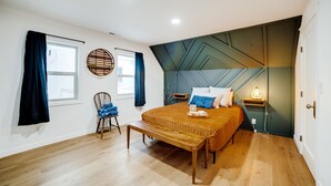 A bedroom with a wooden bed, a bench, a chair, a bedside table with a lamp, and a window with curtains.