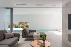 A modern living room with a large glass door leading to a pool area, a sofa, a coffee table with bowls, and a TV on the wall.