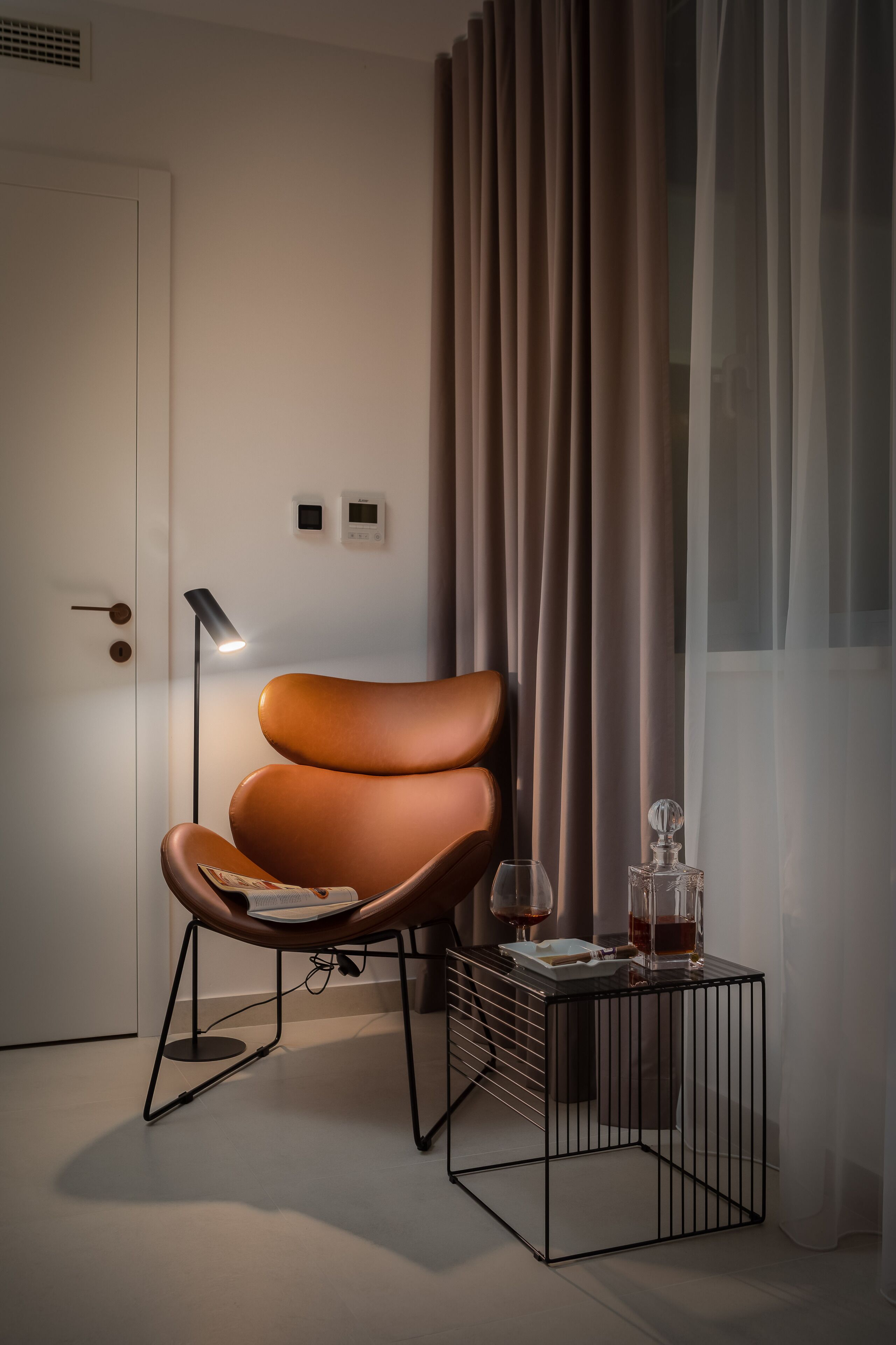 Een moderne kamer met een lederen fauteuil, een klein bijzettafeltje met een lamp, en een deur met sleutelslot.