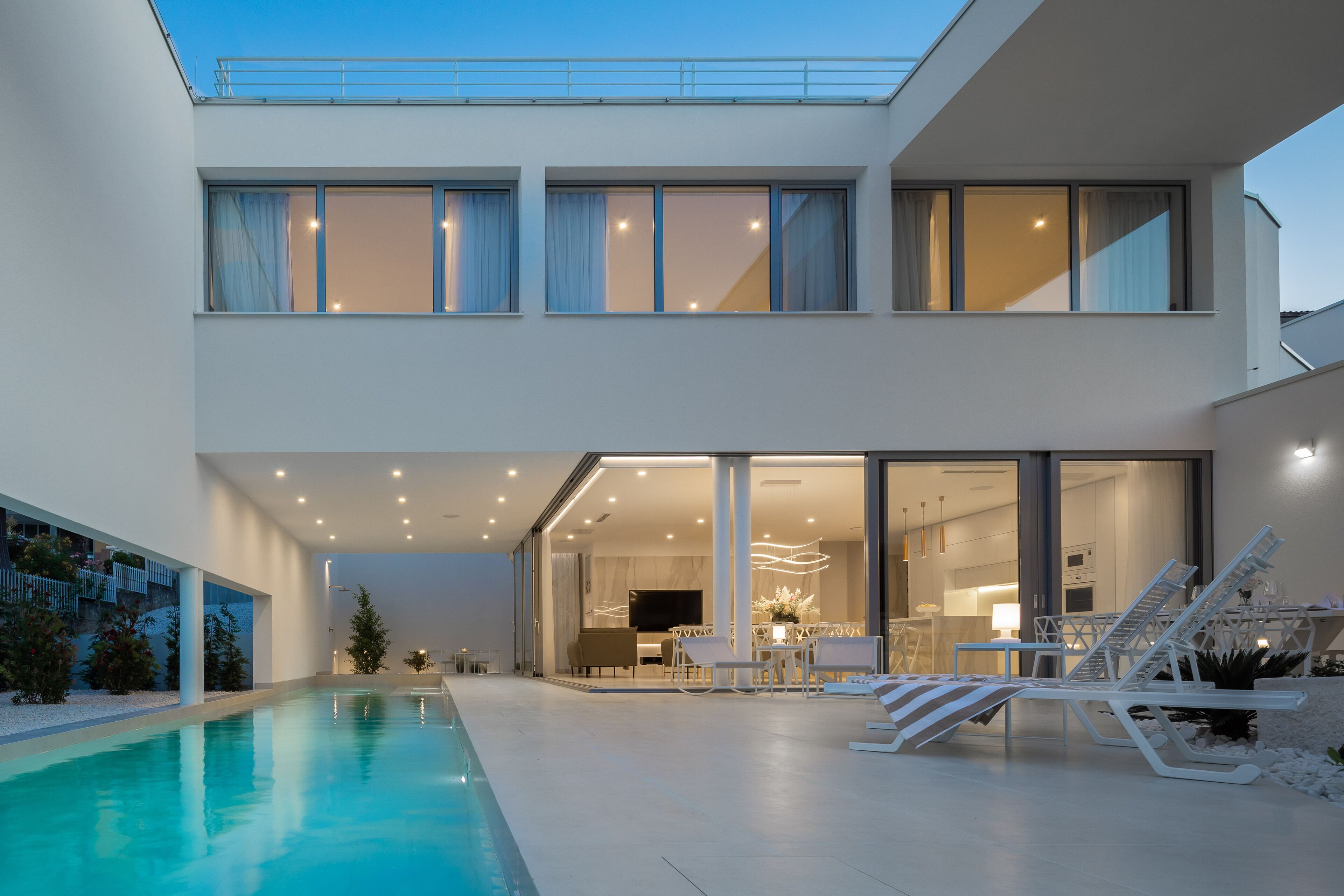 Een modern huis met twee verdiepingen, een zwembad, buitenzitplaatsen en een grote glazen deur die naar de binnenkant leidt.