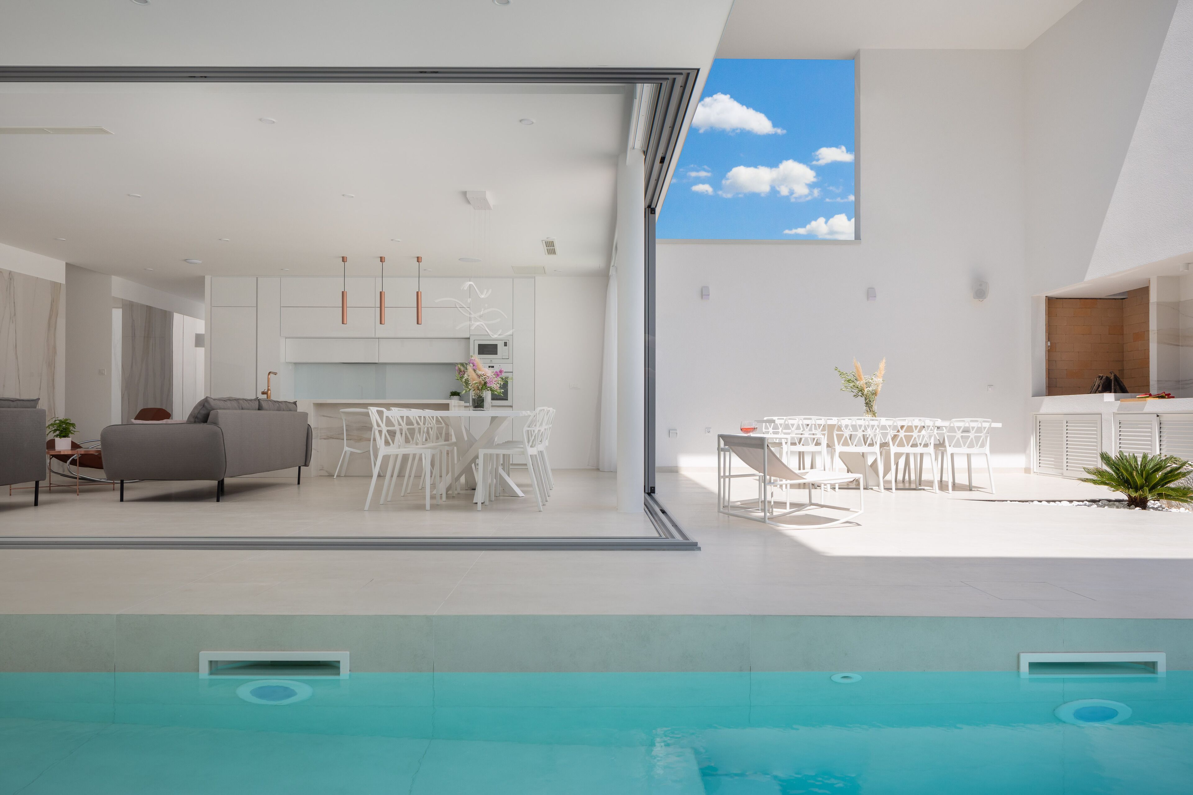 Een modern interieur met een zwembad, witte meubels en uitzicht op een heldere lucht.