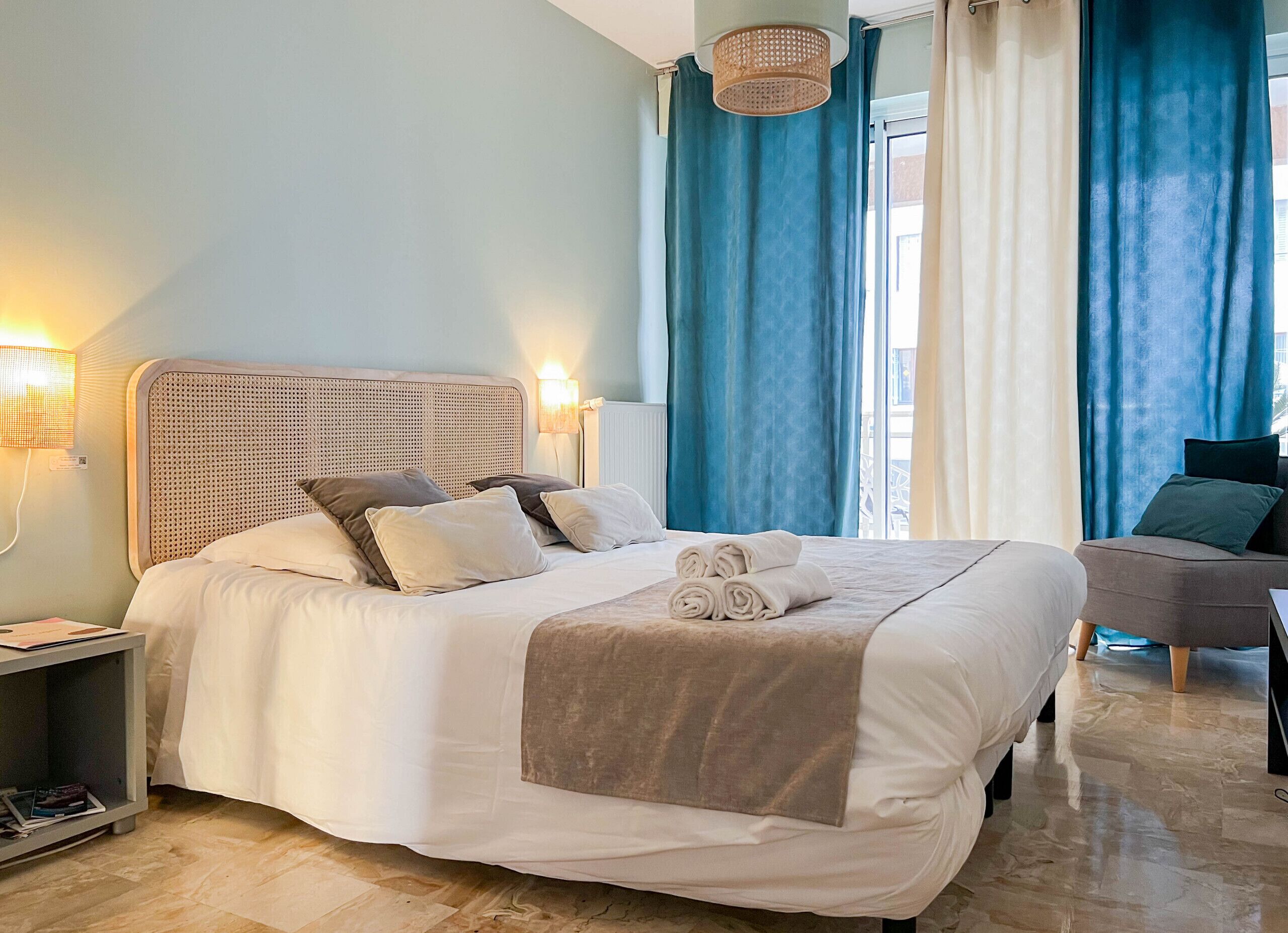 A neatly made bed with pillows and a rolled towel, a bedside table with a lamp, and a chair in a room with blue curtains and a window.