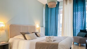 A neatly made bed with pillows and a rolled towel, a bedside table with a lamp, and a chair in a room with blue curtains and a window. - Reve de Jade devant le canal (Annecy)