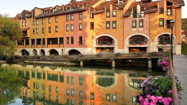 A multi-story hotel with a row of balconies, a reflective water body in front, and a bridge connecting the buildings. - Reve de Jade devant le canal (Annecy)