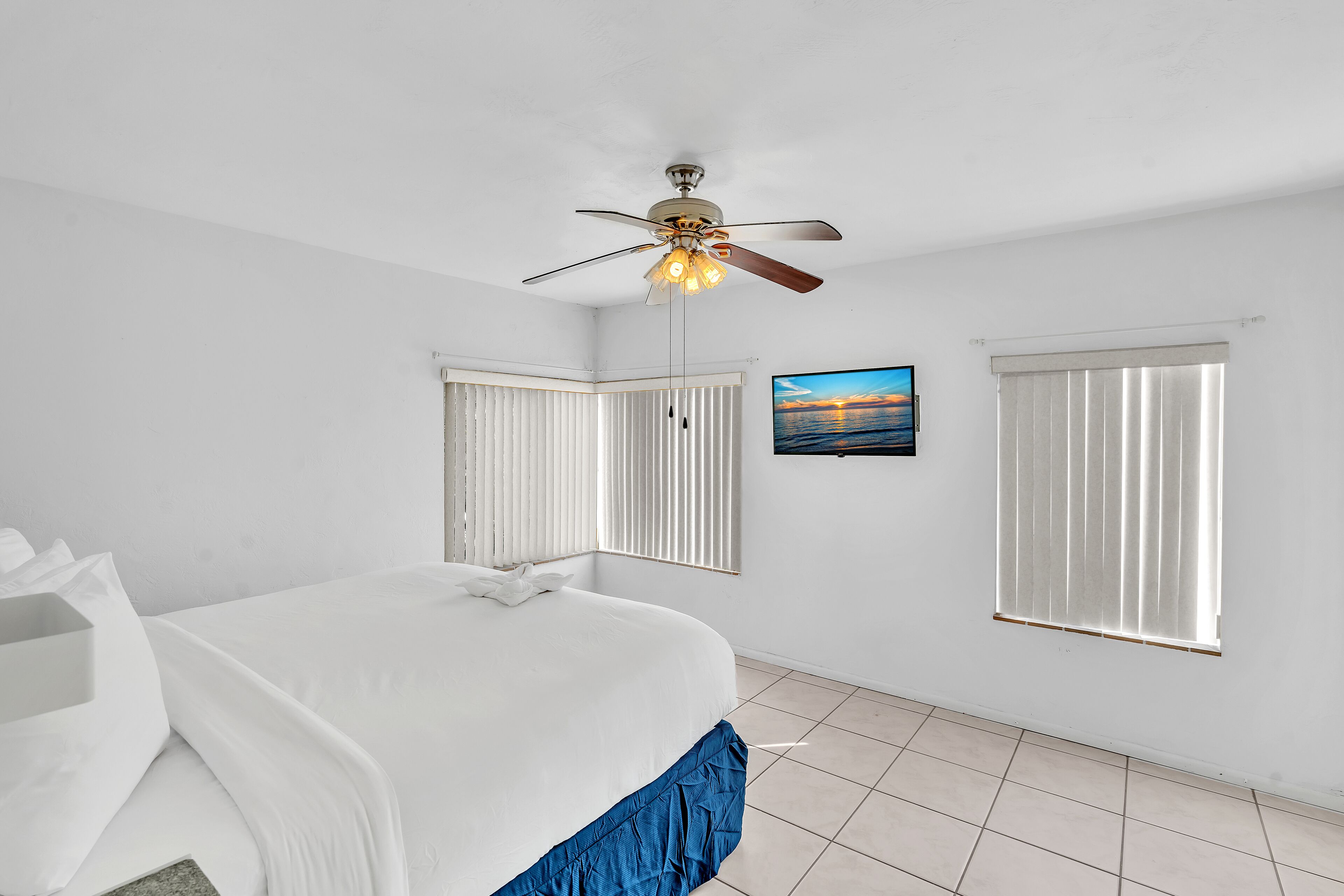 A bedroom with a bed, a ceiling fan, two windows with blinds, and a painting on the wall.