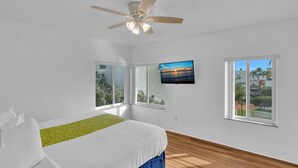Quarto com cama, ventilador de teto, televisão e janelas amplas.
