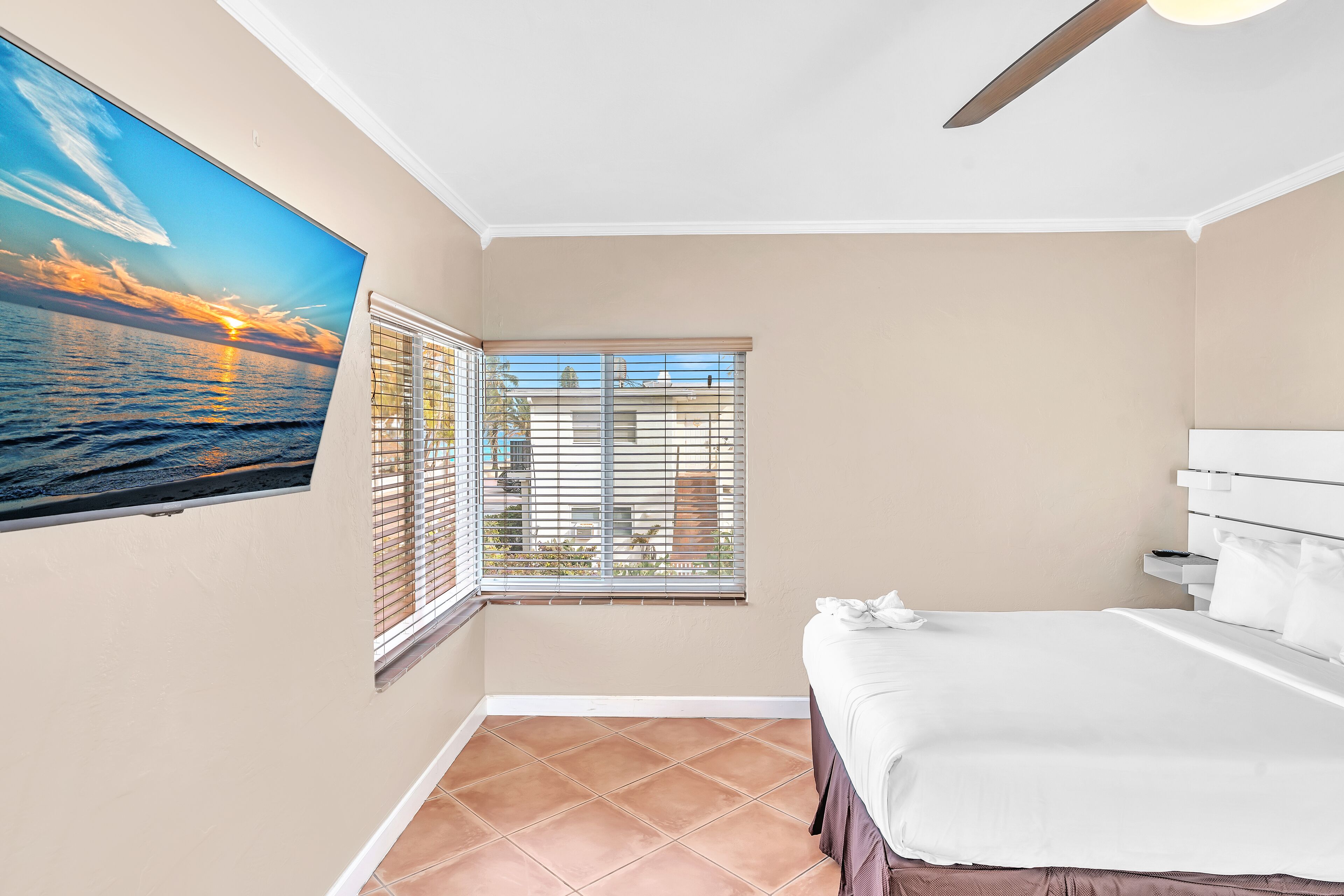 A bedroom with a bed, a window with blinds, a ceiling fan, and a wall-mounted TV displaying a sunset scene.