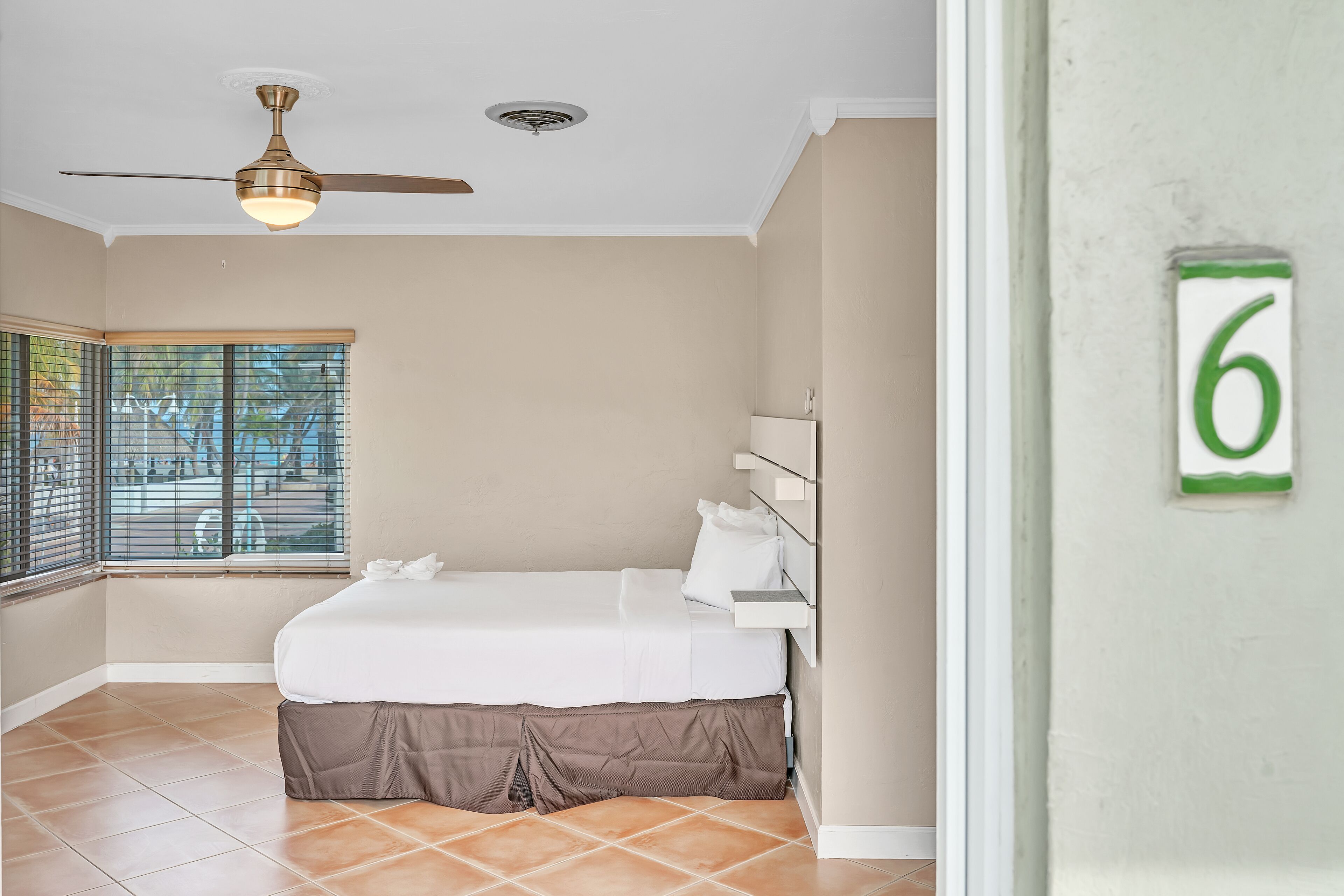 A bedroom with a bed, a ceiling fan, a window with blinds, and a wall-mounted number '6'.