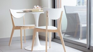 A modern room with two white chairs and a small white table, a window with a view of the outside, and a wicker chair outside the window.