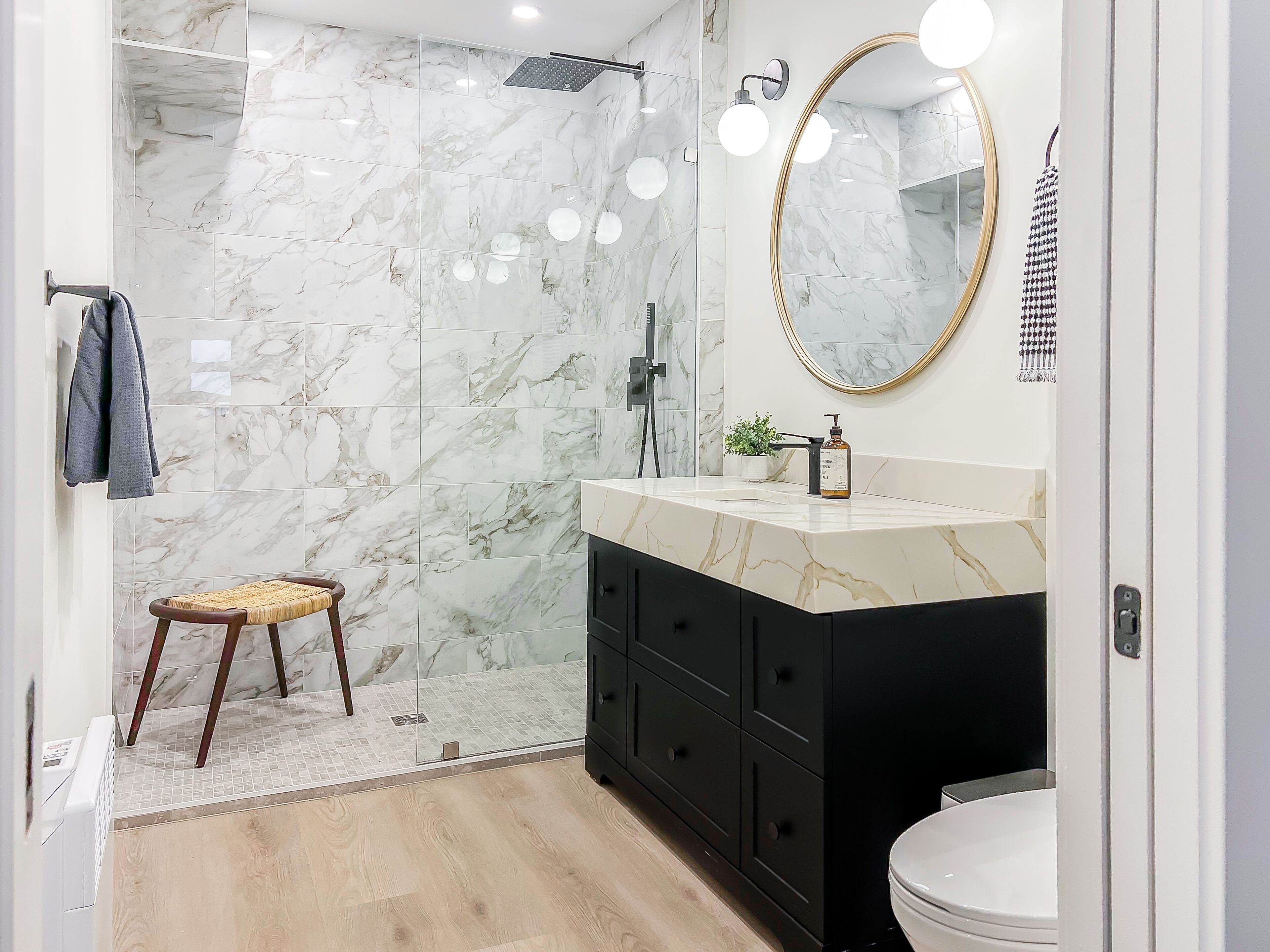 Un baño con ducha de mármol, tocador blanco y negro, espejo redondo y taburete de madera.