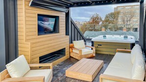 A modern outdoor living area with wooden furniture, a TV mounted on a wooden wall, and a hot tub. - Luxury townhome - Rooftop Patio - Hot Tub (Charlottetown)