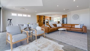 Ein modernes Wohnzimmer mit einem Ecksofa, Sesseln und einem Couchtisch. Im Hintergrund ist eine Küchenzeile mit Holzschränken und einem Kühlschrank zu sehen.