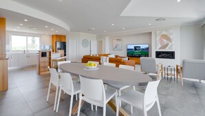 Eine moderne Küche mit weißen Schränken, ein Essbereich mit rundem Tisch und Stühlen sowie ein Wohnbereich mit Sofa und Fernseher.