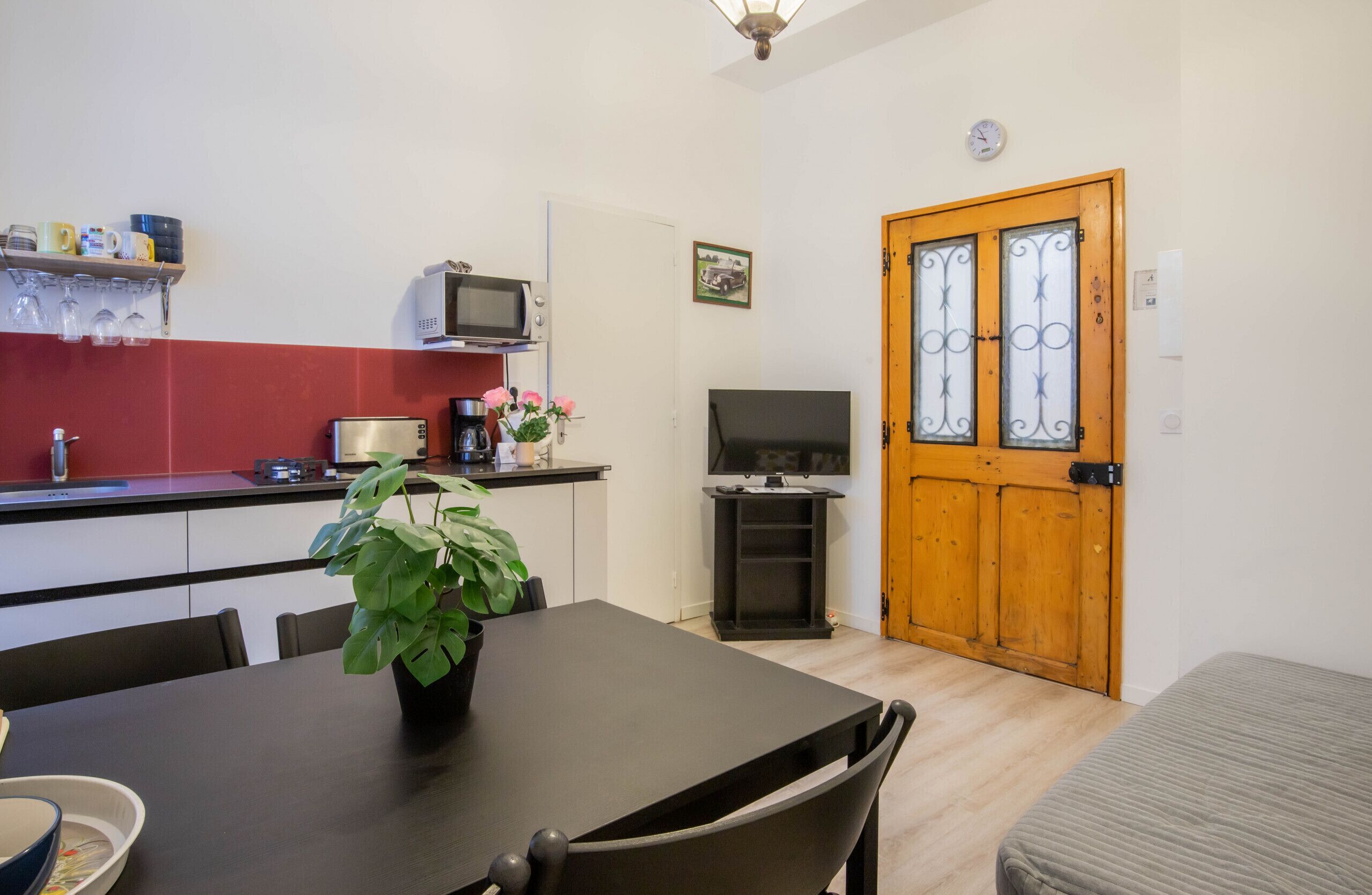A compact kitchen with a microwave, toaster, and a small TV. There's a dining table with chairs and a potted plant. A wooden door with glass panels is visible.