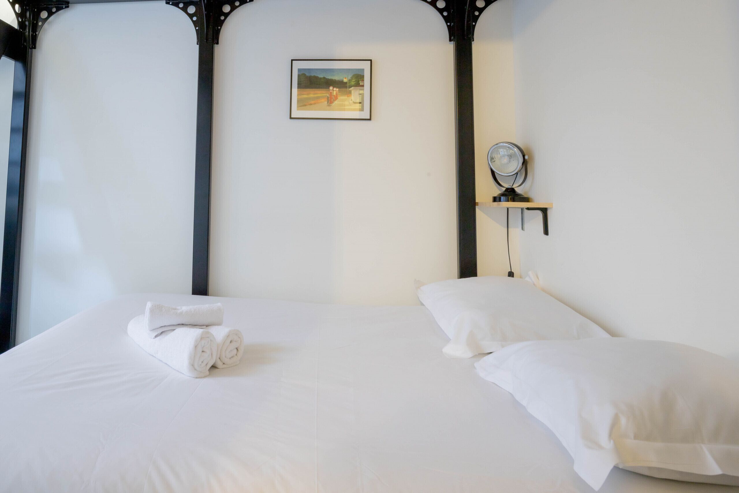 Een net opgemaakt bed met witte lakens en een opgevouwen handdoek, een nachtkastje met een spiegel, en een ingelijst beeld aan de muur.