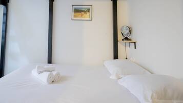 Een net opgemaakt bed met witte lakens en een opgevouwen handdoek, een nachtkastje met een spiegel, en een ingelijst beeld aan de muur.