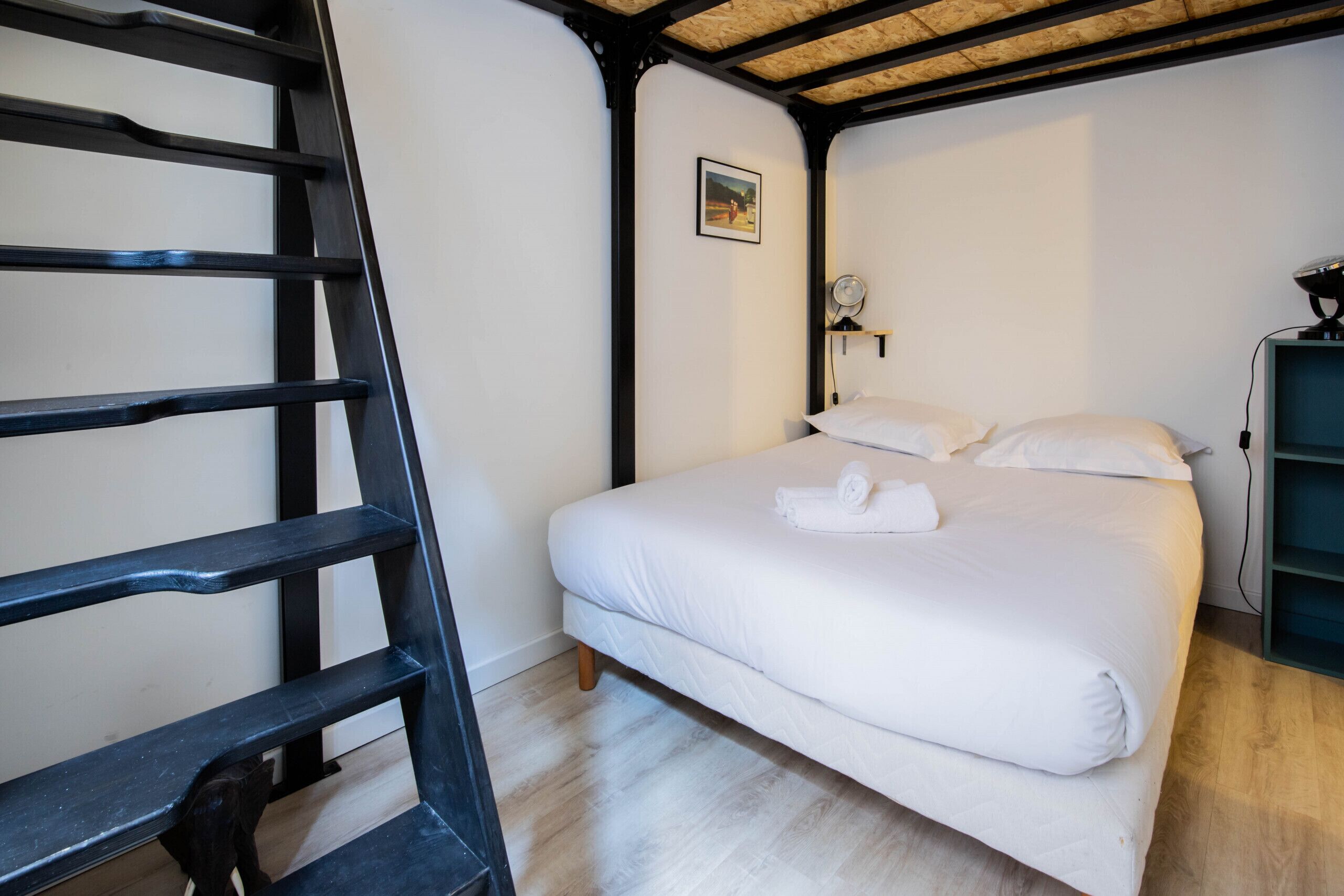 Un dormitorio con una cama, una escalera, un ventilador de pared y un cuadro en la pared.