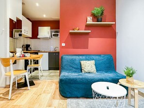 Un espacio habitable compacto con kitchenette, un sofá azul con cojín, una mesa de comedor con sillas y una mesita redonda con una planta.