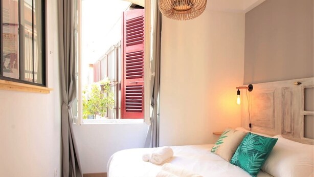Une chambre à coucher avec un lit, une fenêtre avec des rideaux, un luminaire fixé au mur et une vue sur un bâtiment à l’extérieur.