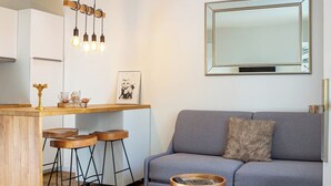 A modern kitchen with a wooden bar, three stools, and pendant lighting. A grey sofa with a throw pillow and a small round table with cutlery are in the living area. - Tahiti Beach 100 m du lac (Annecy)