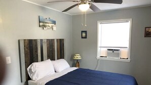 Une chambre à coucher comprenant un lit, un ventilateur de plafond, une fenêtre avec un climatiseur et une table de chevet.