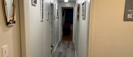 A narrow hallway with light-colored walls and a wooden floor. There are framed pictures on the walls and a sign with text on the right wall.