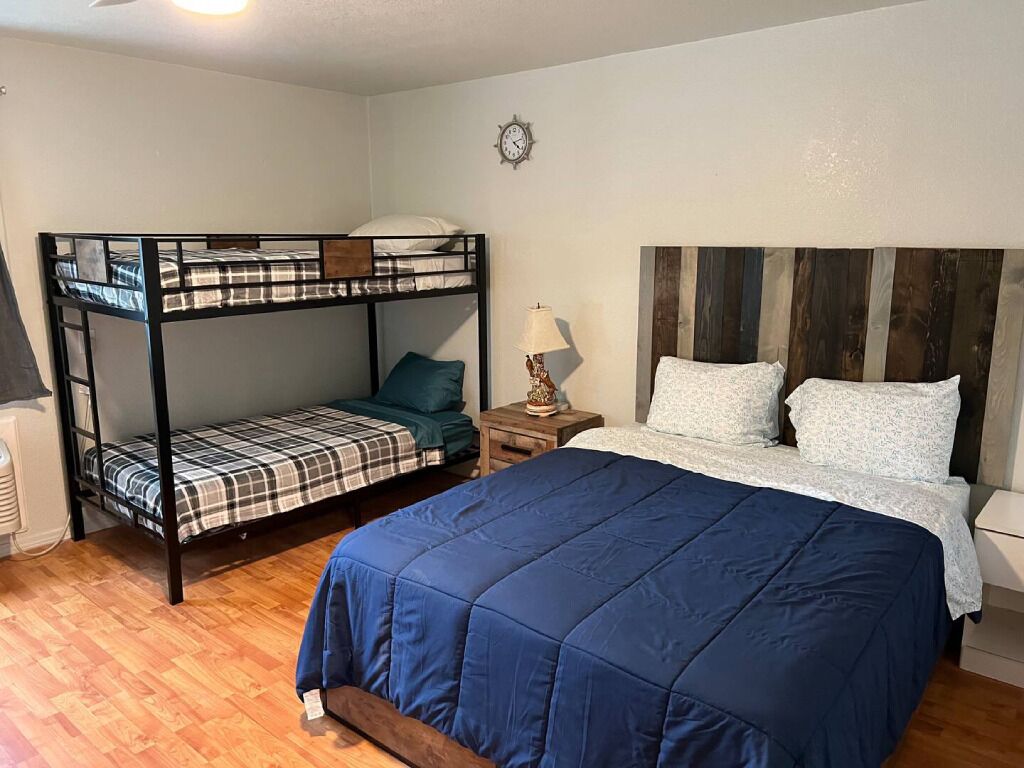 A room with a bunk bed, a single bed, a wooden headboard, a bedside table with a lamp, and a wall clock.