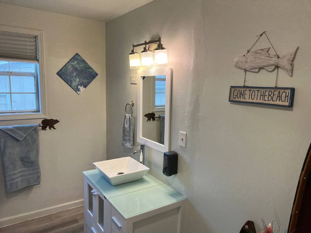 A bathroom with a white sink, a mirror with lights, a towel rack, a wall decoration, and a sign that says "GONE TO THE BEACH".