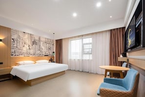Habitación de hotel con cama, zona de estar, televisor montado en la pared y un ventanal amplio.