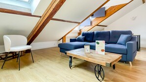 Un dormitorio tipo loft moderno con techo inclinado, vigas de madera, un sofá azul, una silla blanca y una mesa de centro de madera.