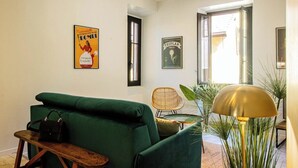 Ein Wohnzimmer mit einem grünen Sofa, einer hölzernen Bank, einem Korbstuhl, einem Tisch mit einer Lampe und zwei Bildern an der Wand.