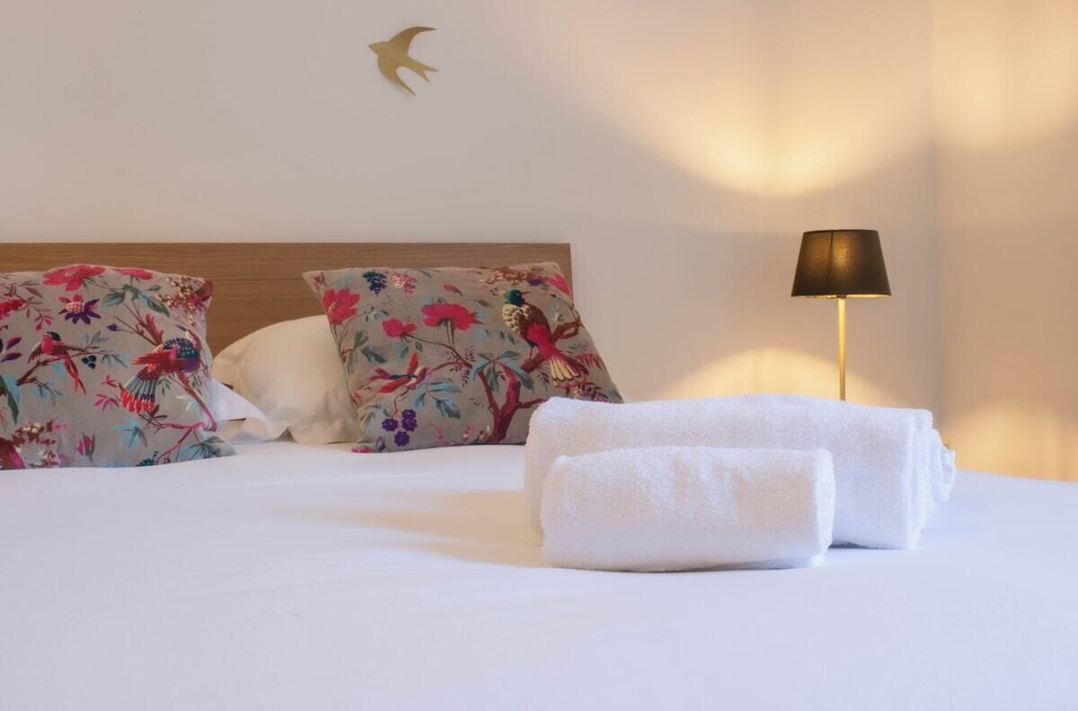 Ein ordentlich bezogenes Bett mit floralen Kissen, einer Lampe und Handtüchern auf einer weißen Bettdecke.