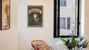 Ein Zimmer mit Korbsessel, einem Poster an der Wand und Blick auf eine Straße durch ein Fenster.