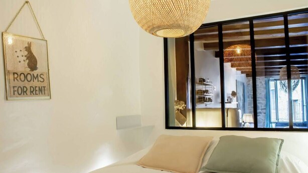 Un dormitorio con una cama, dos almohadas, una lámpara colgante y una ventana que da a una habitación con una silla y una mesa.
