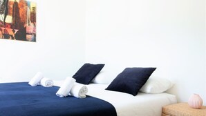 A hotel room with a bed, pillows, a rolled towel, a small wicker side table with a vase, and a painting on the wall. - Garden plazza T2 standing parking (Annecy)