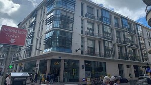 A multi-story building with large windows and balconies, a ticket booth sign, and a pedestrian area with people and bicycles. - Garden plazza T2 standing parking (Annecy)