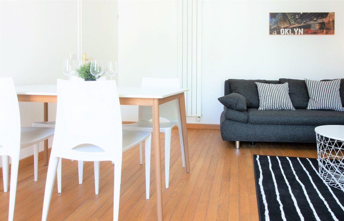 Een moderne woonkamer met een grijze bank, witte stoelen, een houten eettafel en een zwart-wit gestreept tapijt.