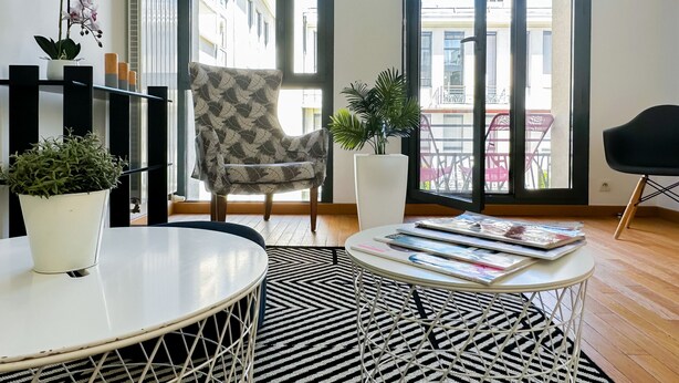 A modern living room with a patterned armchair, a round table with magazines, a potted plant, and a view of the outside balcony. - Garden plazza T2 standing parking (Annecy)