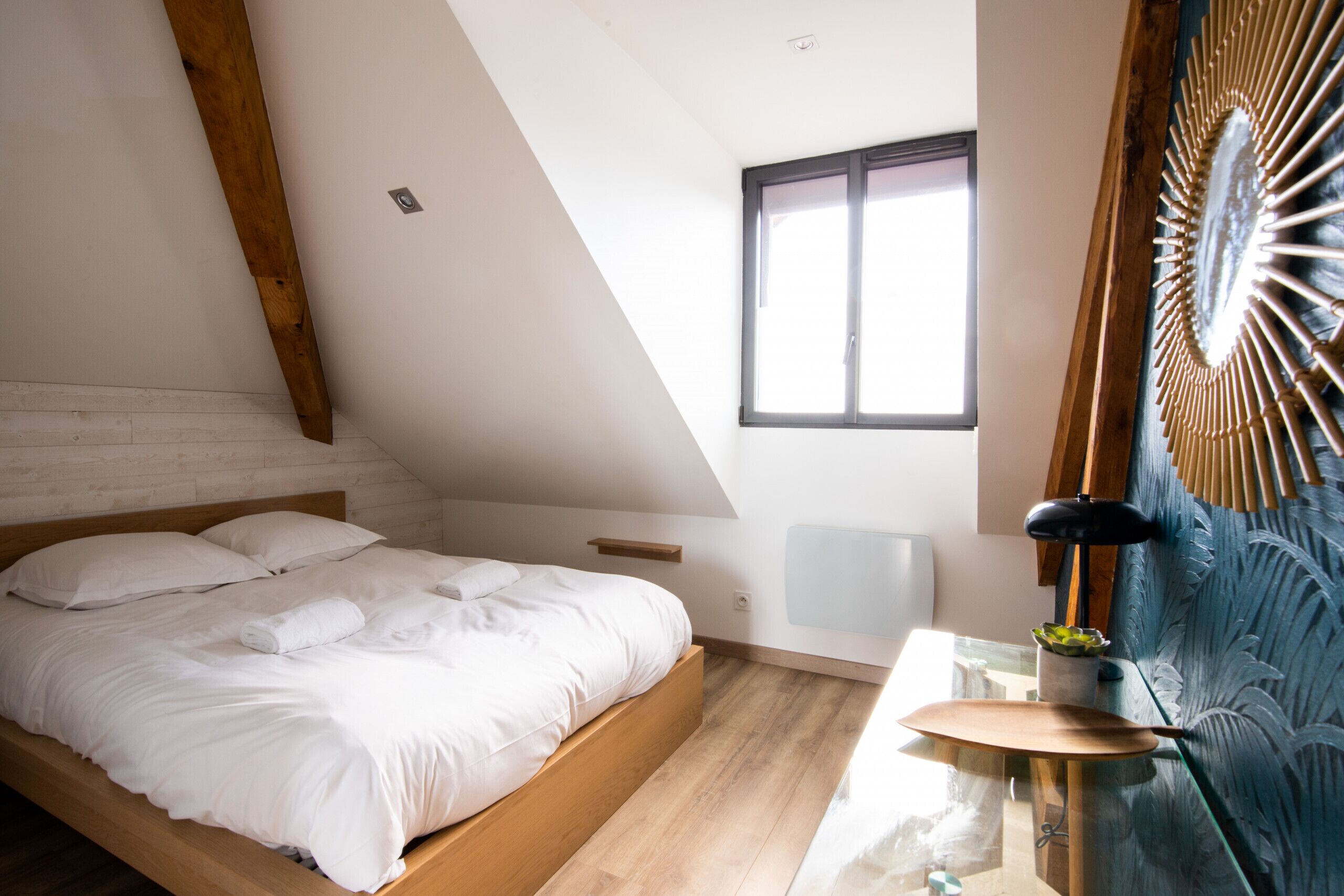 Een slaapkamer met een schuine plafond, een bed met witte lakens, een tafeltje met een lamp en een decoratief wandobject.