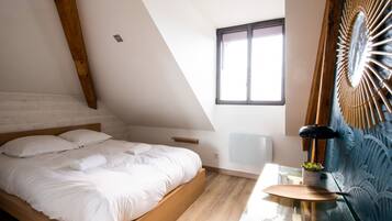Een slaapkamer met een schuine plafond, een bed met witte lakens, een tafeltje met een lamp en een decoratief wandobject.