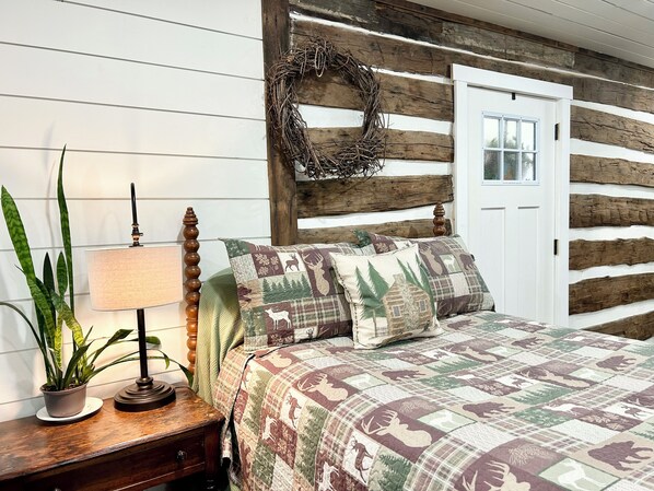 A cozy bedroom with a wooden log wall, a bed with a quilt featuring animal prints, a nightstand with a lamp, and a potted plant.